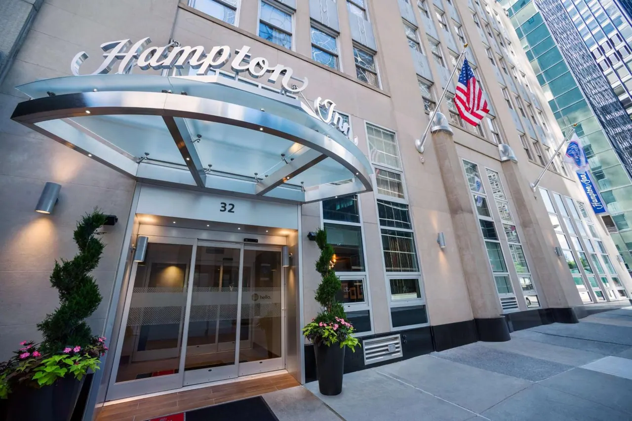Street-level entrance of a Hampton Inn hotel with a glass canopy, two flags, topiary planters and a revolving-style glass door under the building sign.