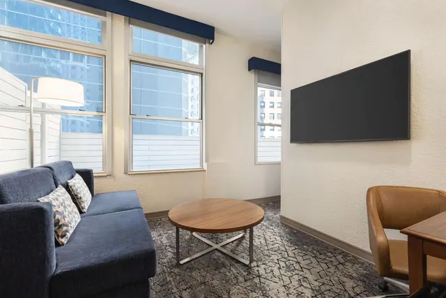 A living area with a navy sofa, a round wooden coffee table, a wall-mounted television and a tan leather chair, with windows looking onto city buildings.
