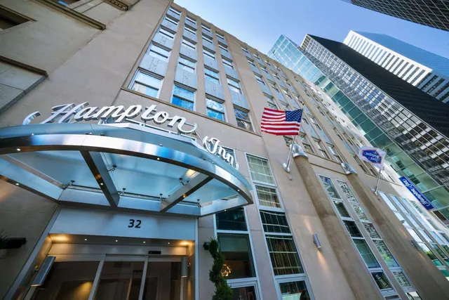 Upward view of the Hampton Inn facade with its glass entrance canopy, a street number above the doors, an American flag on the wall, and neighbouring glass skyscrapers under a clear blue sky.