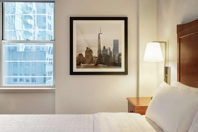 A bedroom with a made bed, a wall lamp above a wooden nightstand, a framed black-and-white city skyline print, and a window looking out on neighbouring buildings.