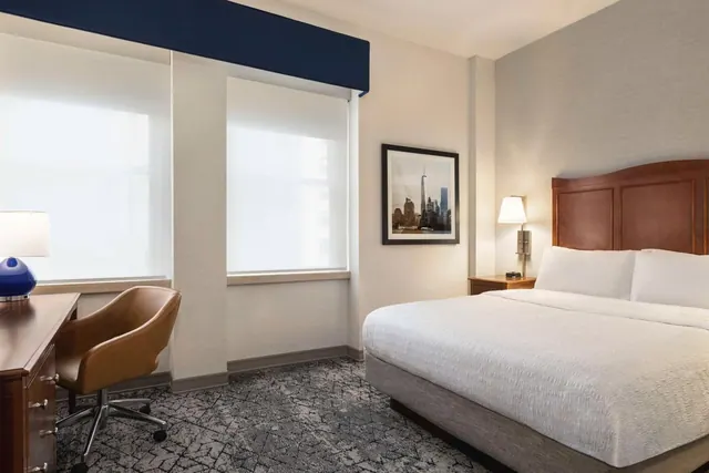 A king bed with white linens and a wooden headboard sits beside a wooden desk with a brown leather chair, and a framed Manhattan skyline print hangs on the wall.