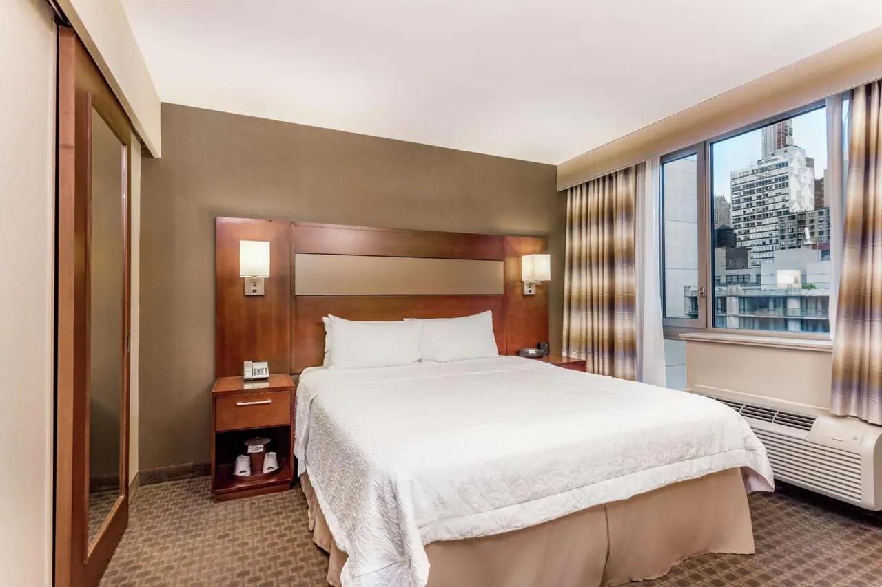 A king bed with a wooden headboard sits beside a window overlooking city buildings in a hotel bedroom.