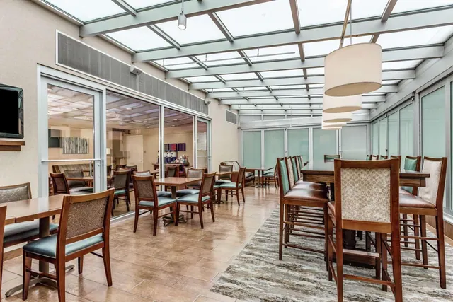 A dining room with a glass skylight ceiling, wooden chairs with teal seats at square and high tables, pendant lamps, and a wall-mounted television.