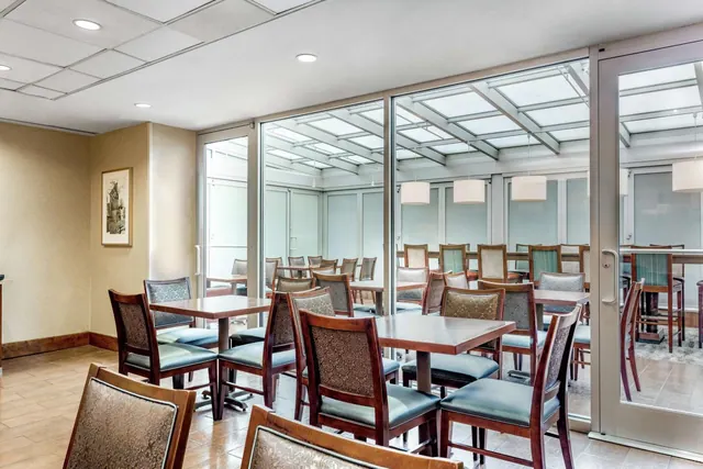 A dining room with wooden tables and upholstered chairs beside floor-to-ceiling glass walls that look onto an enclosed glass-roofed courtyard.