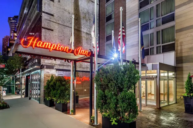 Exterior of a Hampton Inn hotel entrance at dusk, with a lit red neon sign, flagpoles, potted evergreens and glass entrance doors.
