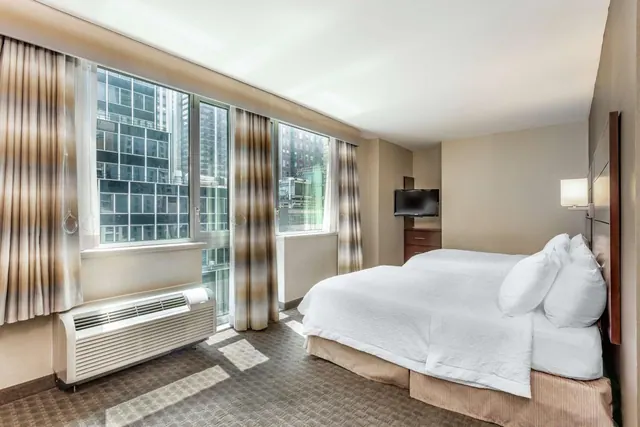 A bedroom with a made double bed, a wall-mounted television, a wall air conditioning unit, and large windows looking out onto city buildings.