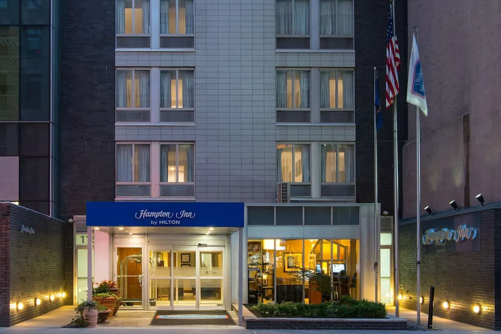Entrance of a Hampton Inn by Hilton building at dusk, with a blue canopy, lit windows, and American and other flags on poles.