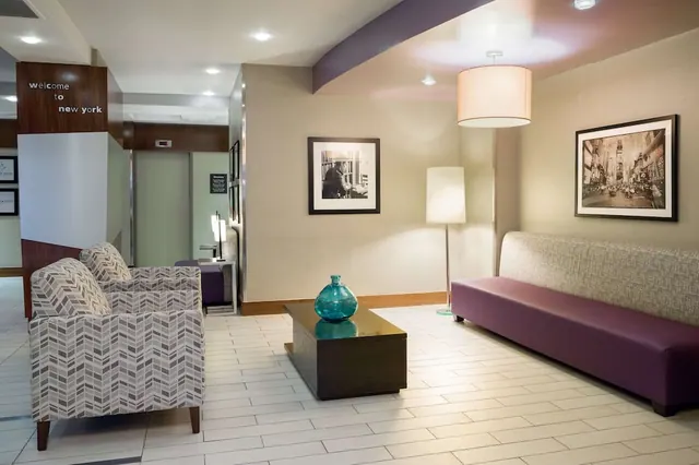 A hotel lobby seating area with chevron-patterned armchairs, a purple bench, a teal glass vase on a dark coffee table, and a wall sign reading welcome to new york.