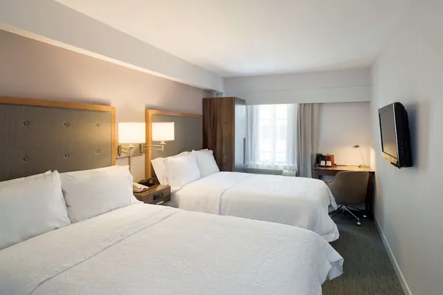 A hotel room with two double beds, a wall-mounted television, a desk with a chair, and a window with curtains.
