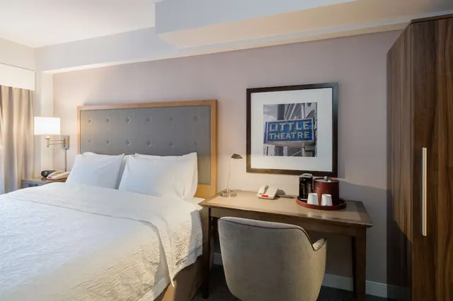 A hotel bedroom with a king bed under white linens, a wooden desk with a grey chair and a coffee station, a tall wooden wardrobe, and a framed print of a Little Theatre sign on the wall.