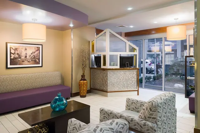 A hotel lobby with a reception desk, a purple bench, patterned armchairs and a coffee table, with glass doors looking out onto a street and a parked car.