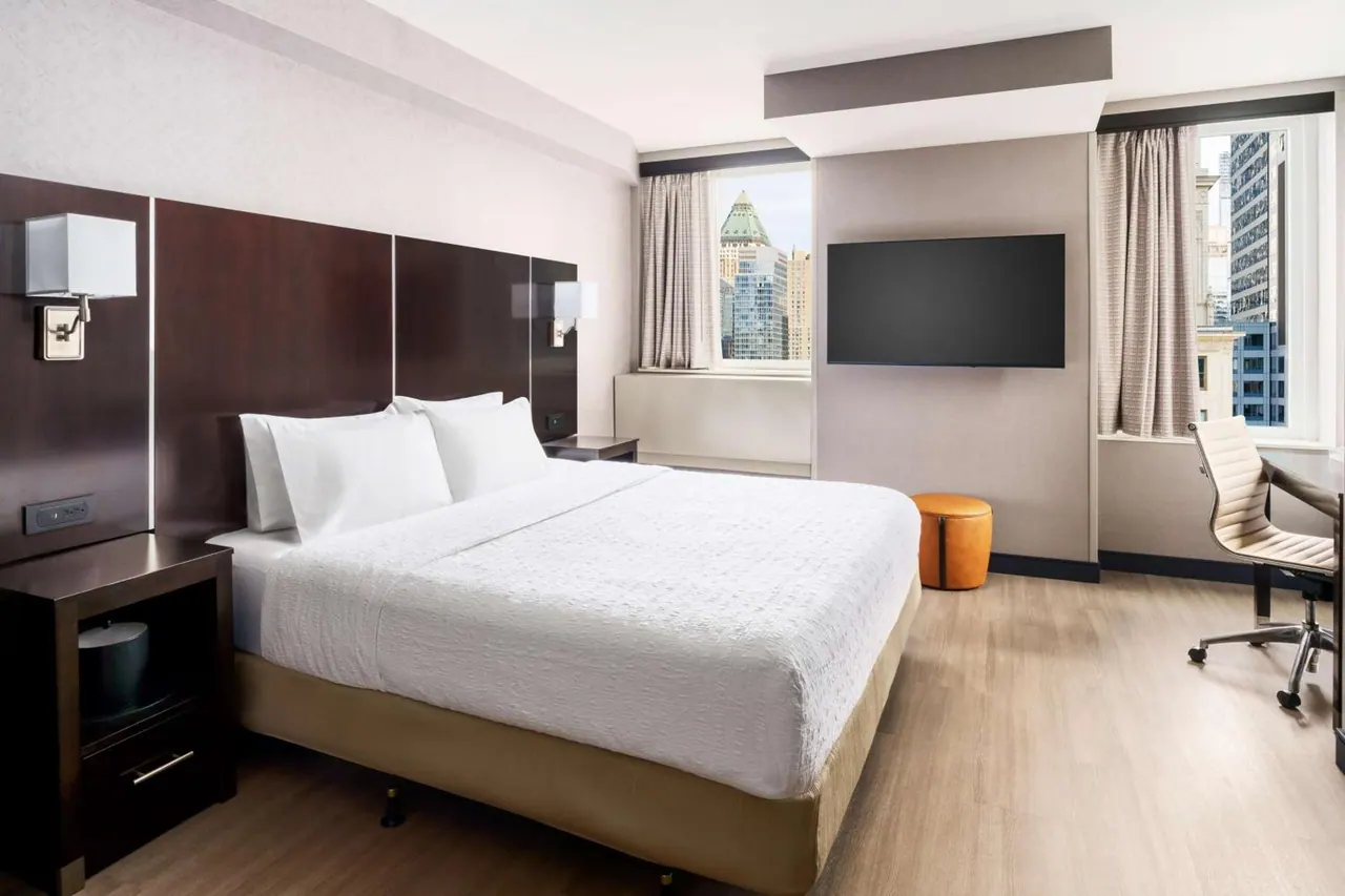 A bedroom with a king bed, a wall-mounted television, an orange ottoman, and a desk with a chair, with windows looking out onto city buildings.