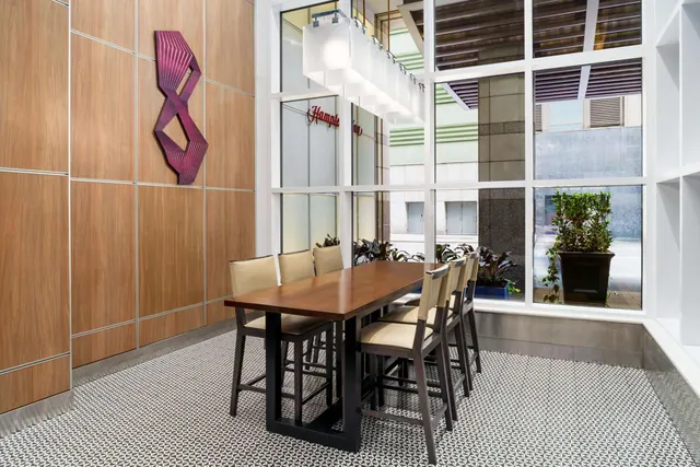 A lobby seating area with a long wooden high table, tall upholstered chairs, a wood-panelled wall with a magenta metal sculpture, and large windows onto a street.