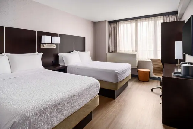 A hotel room with two queen beds with white bedding, a dark wood desk with a chair, a wall-mounted television, and a window with sheer curtains.