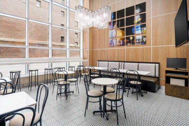 A dining area with round tables, dark bentwood-style chairs, a cushioned banquette, a wall-mounted television, and tall windows facing a brick building.