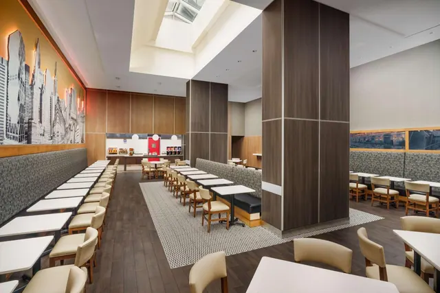 A bright dining room with white tables, beige chairs, a upholstered banquette, a city skyline mural on the wall and a skylight above.
