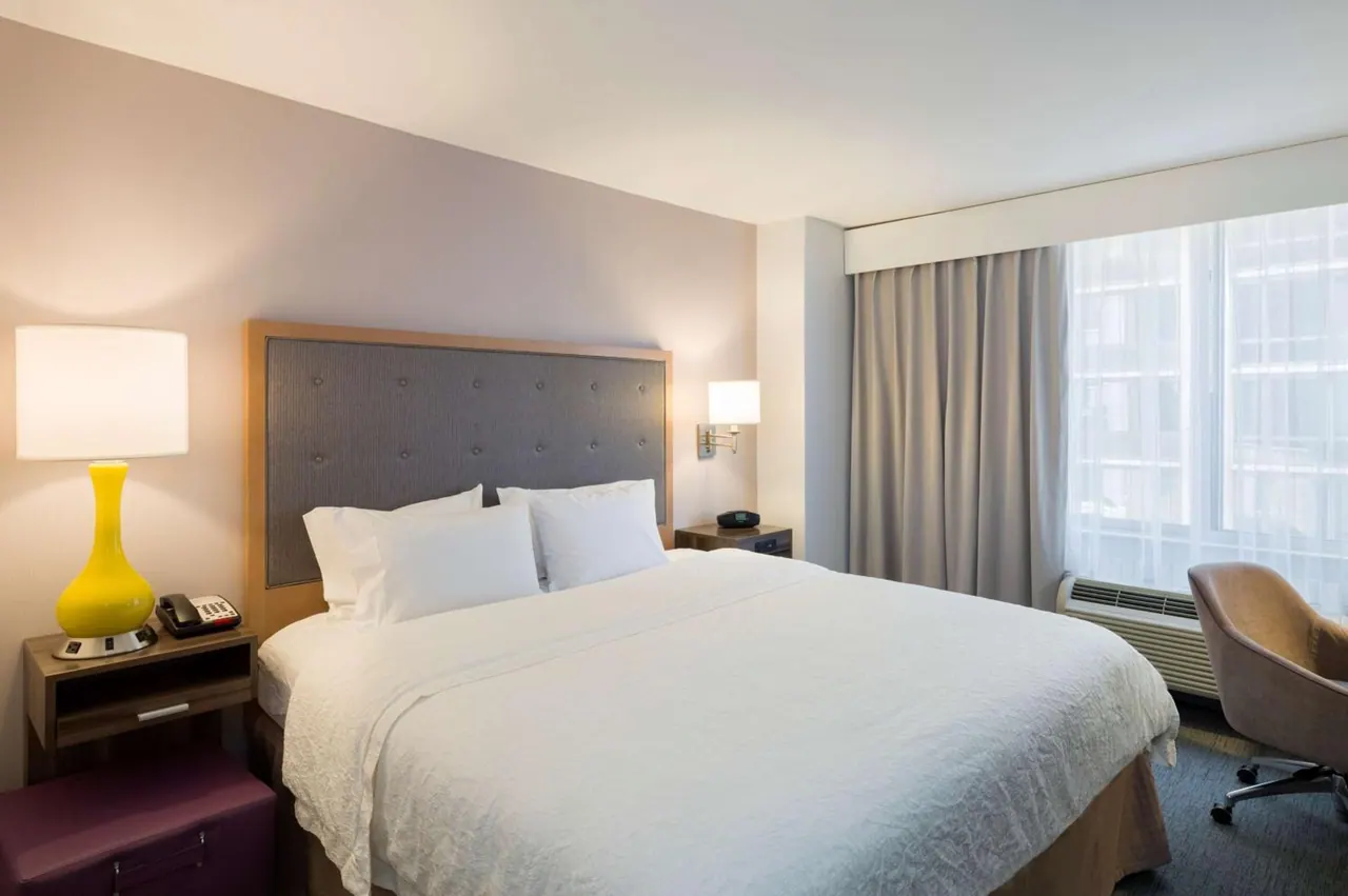 A king bed with white linens sits between two bedside tables with lamps, beside a window with sheer curtains and a wall-mounted air conditioner unit.