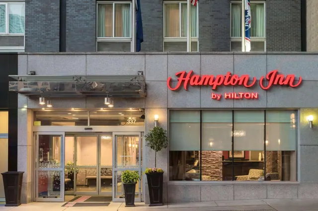 Street-level exterior of a Hampton Inn by Hilton building with illuminated red signage, a glass-fronted entrance, and potted plants by the door.