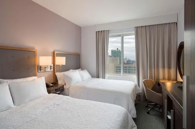 A bedroom with two white-covered double beds, a window with a view over city buildings, and a desk with a chair beside a wooden cabinet.
