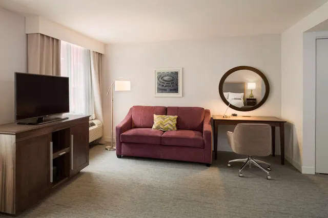 A living area with a red sofa, a television on a wooden cabinet, a desk with a round mirror, and a grey upholstered chair.