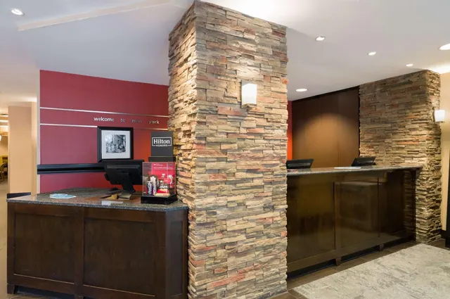 The lobby reception area has two dark wood front desks, a tall stone-clad pillar, and a red wall with a 'welcome to new york' sign.