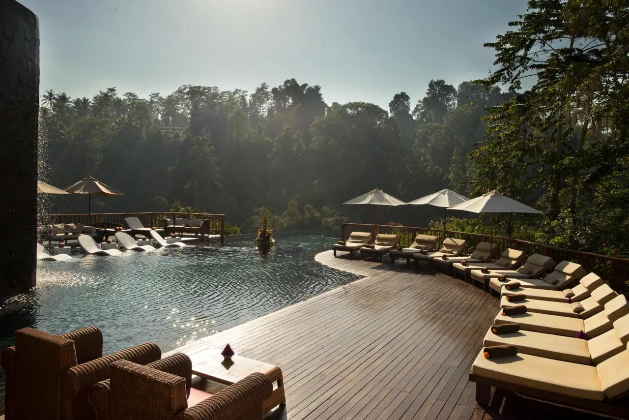 An infinity pool with cream sun loungers and parasol umbrellas on a timber deck, overlooking dense green jungle under hazy light.