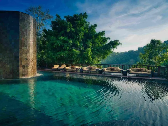 A tiled water feature stands beside a turquoise pool, with cushioned daybeds along the deck and jungle trees and hills behind.
