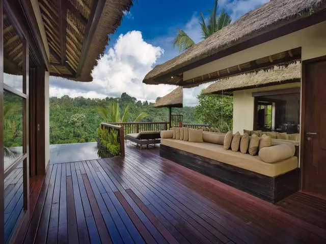 A covered wooden terrace with a long cushioned daybed, a lounger, and an infinity pool edge overlooking forested hills under a partly cloudy sky.