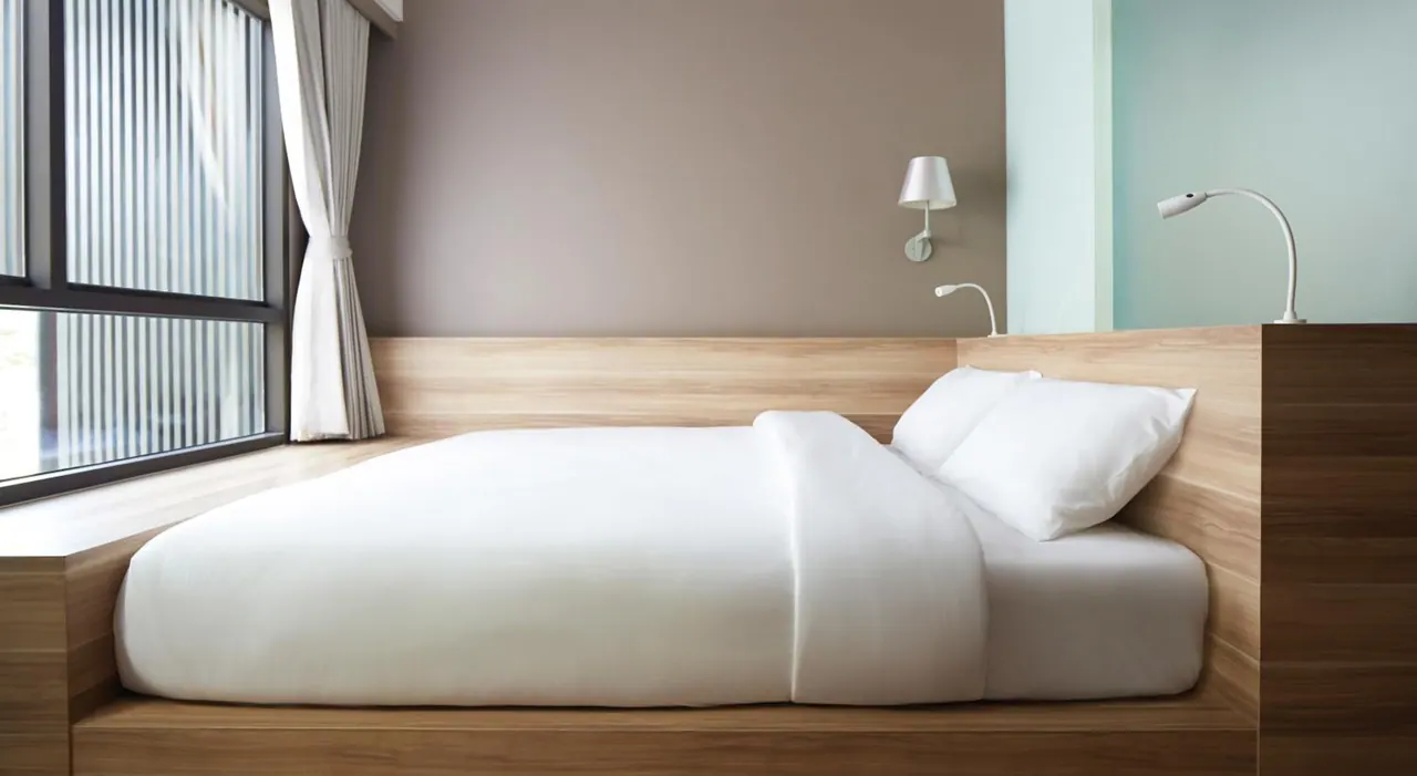 A double bed with white linens, a wooden platform frame and wall-mounted lamps sits beside a window with side curtains.