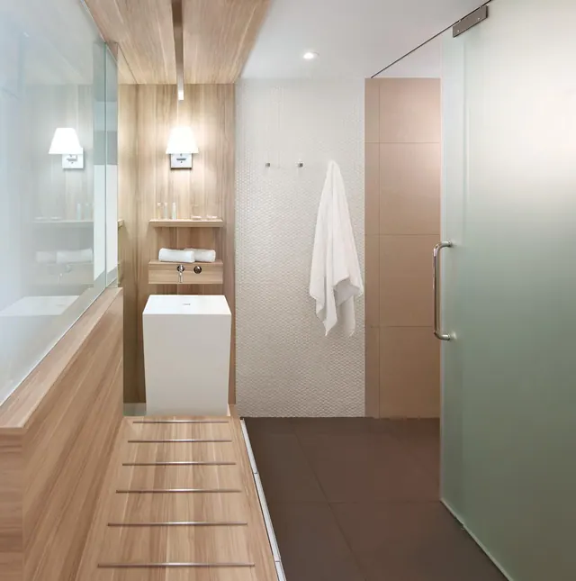 A bathroom with a freestanding white pedestal basin, a wood-panelled wall, a frosted glass shower enclosure, and a bathtub visible behind glass on the left.
