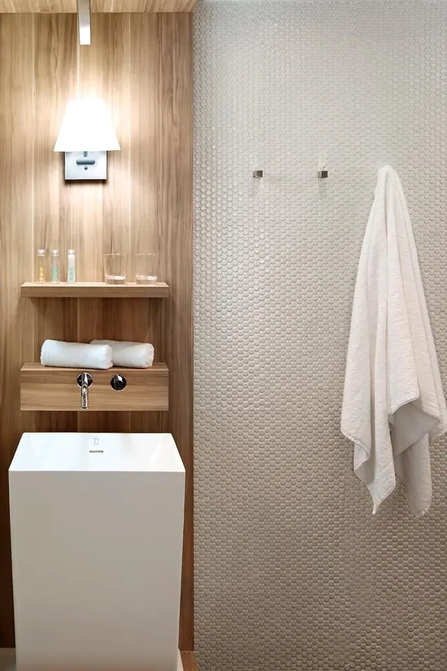 A bathroom with a wood-panelled wall, a wall lamp, a shelf holding small toiletry bottles and glasses, rolled white towels, and a freestanding white basin in front of a textured tiled wall with a hanging towel.