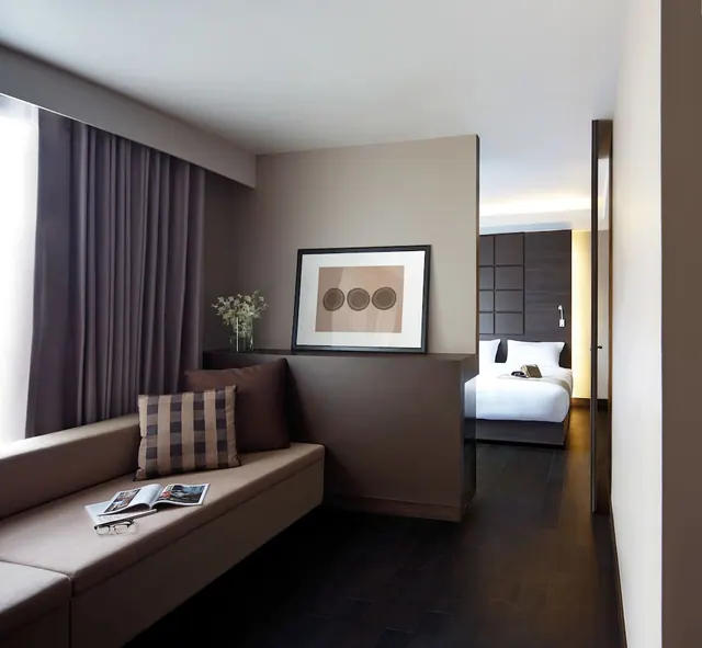 A hotel room with a built-in bench seat by the curtained window, cushions, an open magazine and glasses, and a bed visible through the doorway.