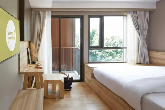 A bedroom with a large bed, a wooden desk holding a camera and glasses, a wall-mounted TV, and a sliding door to a balcony, with trees visible through the window.
