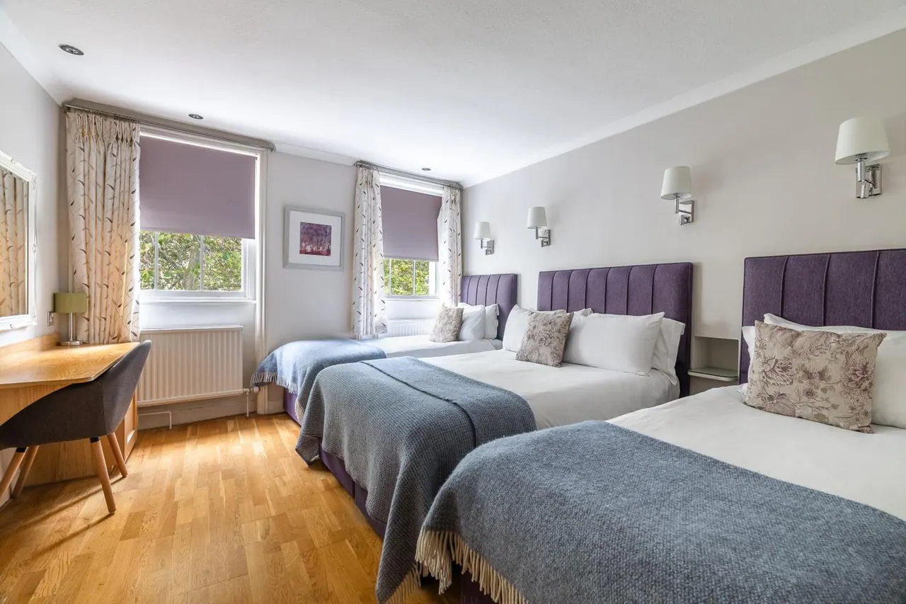 A bedroom with three beds with purple upholstered headboards, a wooden desk and chair, and windows with purple blinds looking out onto trees.