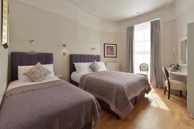 Two single beds with purple headboards and throws stand in a bedroom with a wooden floor, a dressing table with a chair, and a tall window with grey curtains.