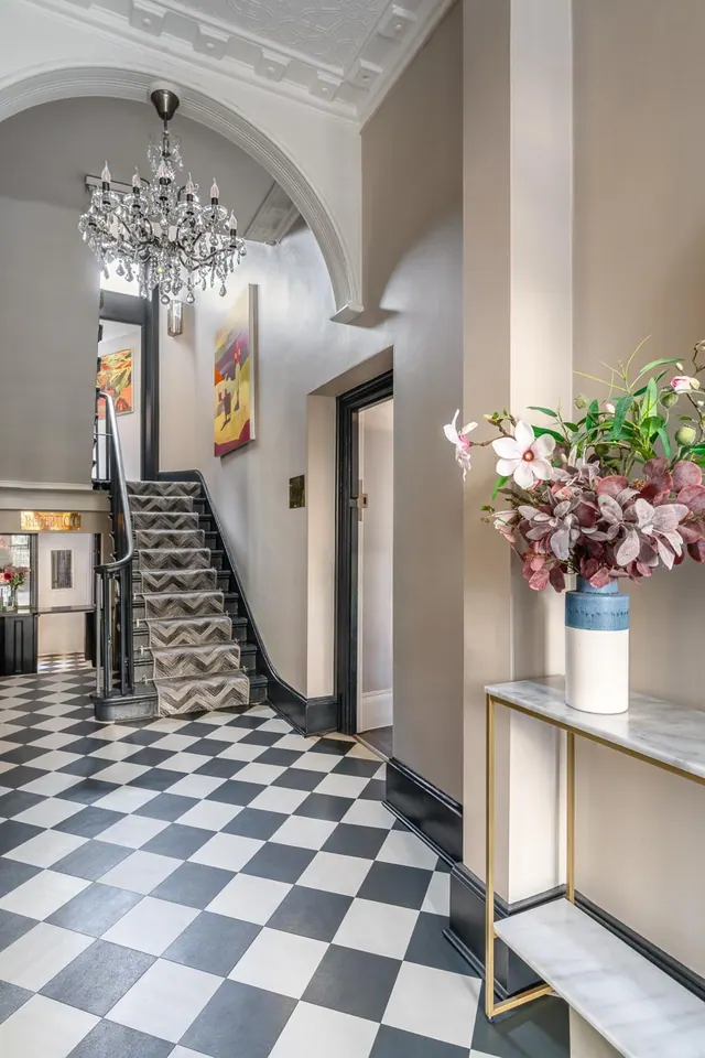 A lobby with a black and white checkerboard floor, a crystal chandelier under an arched ceiling, a patterned carpeted staircase and a flower arrangement on a marble console table.