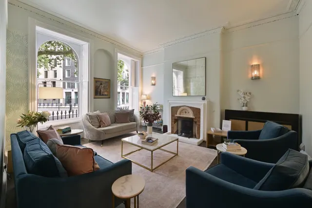 A staged living room with blue armchairs, a grey sofa, a fireplace and tall sash windows looking onto a street.