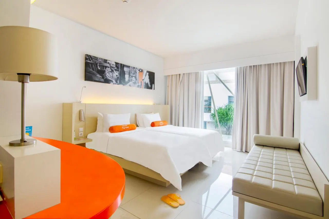 A hotel room with two white beds with orange bolsters, an orange curved desk, a cream bench, and a glass door looking onto a courtyard with a tree.