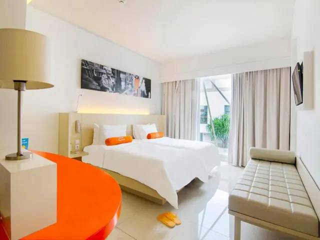 A bedroom with a large white bed with orange bolsters, a leather bench at the foot of the bed, a wall-mounted television, and a glass door with curtains opening onto a plant-filled courtyard.