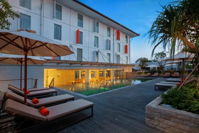 Outdoor pool with sun loungers and shade umbrellas on a timber deck beside a white hotel building at dusk.