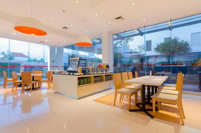 A breakfast restaurant with orange pendant lamps, a buffet counter with drink dispensers and plates, wooden chairs at tables, and floor-to-ceiling glass walls looking onto a courtyard with trees.