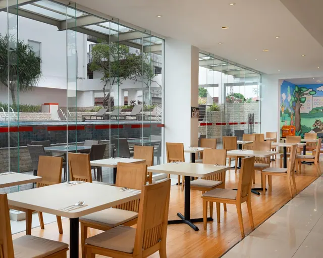 A dining room with wooden chairs and white tables beside floor-to-ceiling glass walls that overlook a pool with sun loungers, and a children's mural on the right wall.