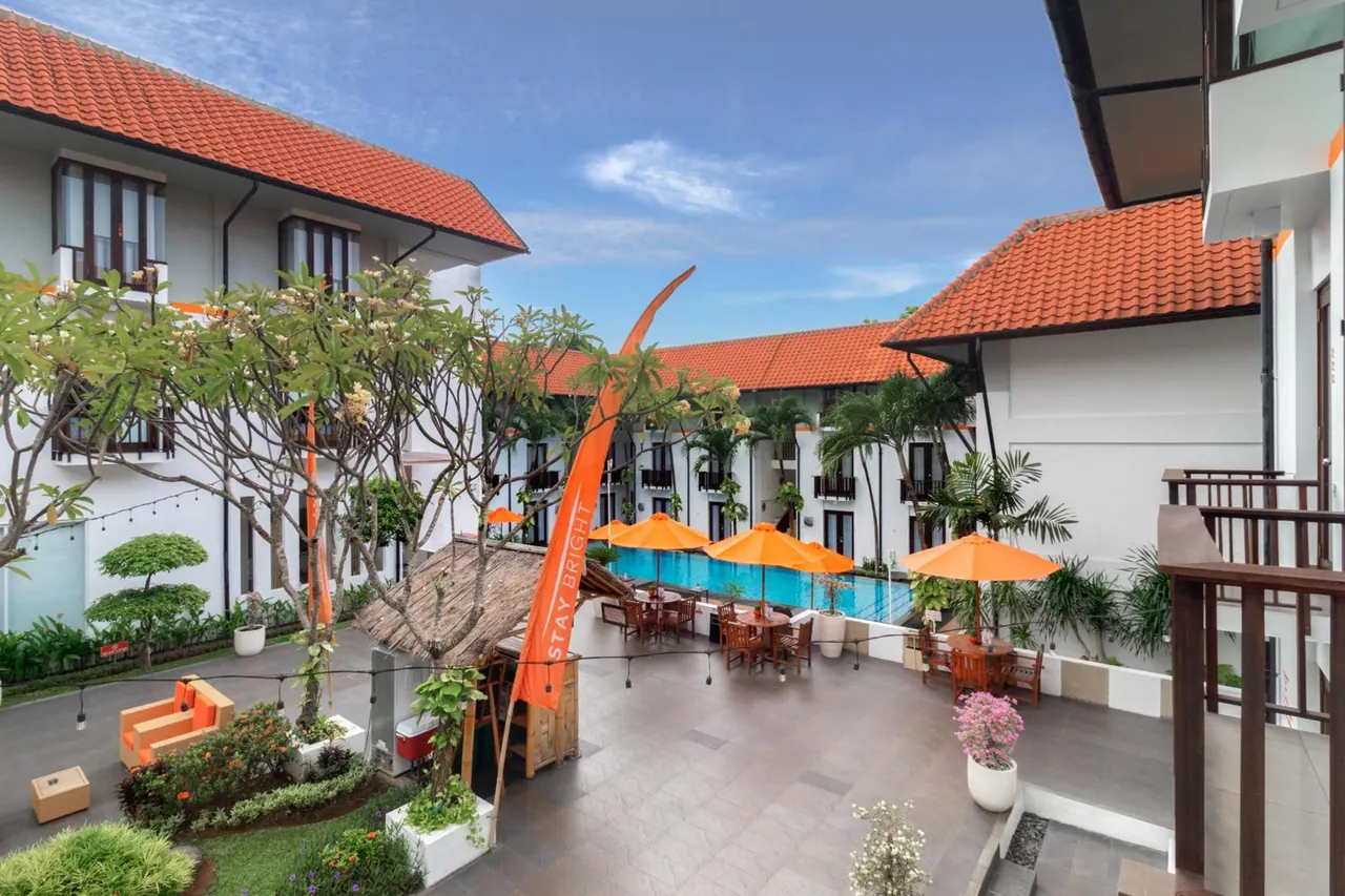 A rectangular outdoor swimming pool in a courtyard, with orange parasols and dining tables beside it and white two-storey buildings with terracotta roofs around it.