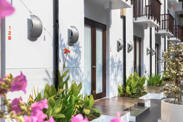 Exterior of a white multi-level guest room block with dark timber doors, upper balconies, room numbers 128 and 130, and tropical plants along a paved walkway.