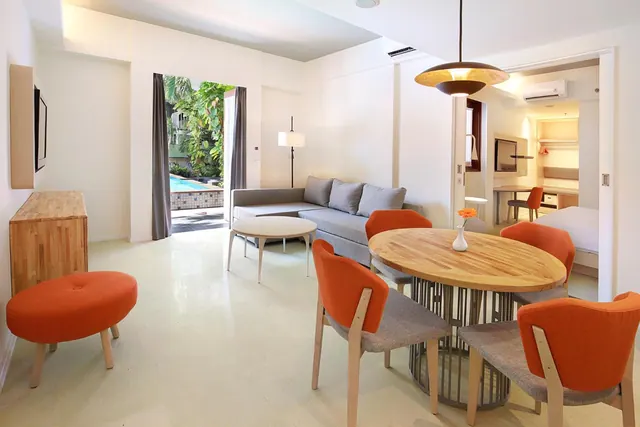 A living area with a grey sofa, a round wooden dining table with orange chairs, and an open doorway onto a pool and green plants.