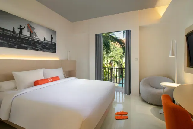 A bedroom with a large white bed, a grey armchair, a desk with an orange chair, a wall-mounted television, and an open door to a balcony with palm trees outside.