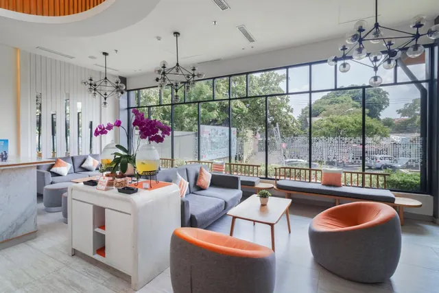 A lobby lounge with grey sofas, orange armchairs, a white island table with an orchid arrangement, and floor-to-ceiling windows looking onto a street with trees and parked cars.