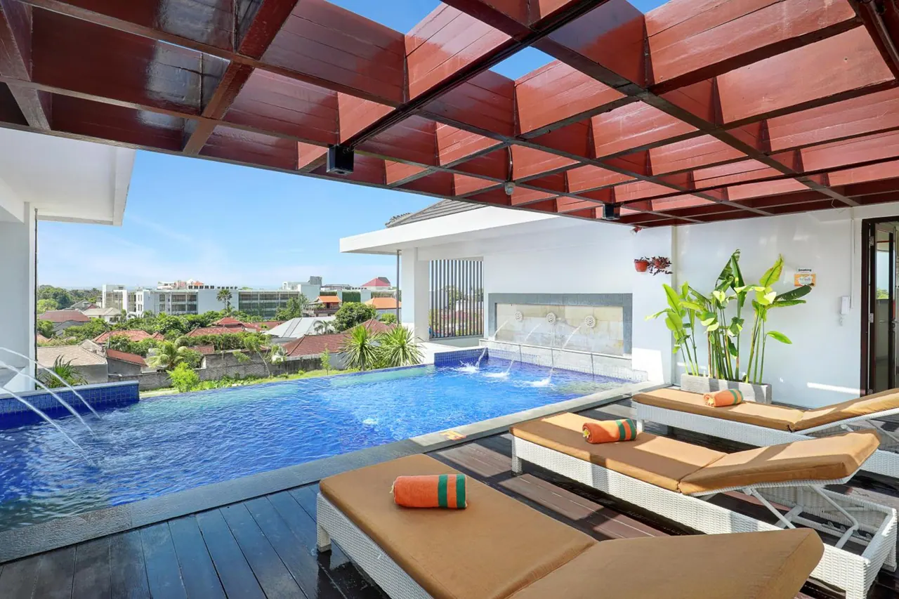A rooftop pool sits beside white sun loungers with brown cushions under a wooden pergola, with nearby buildings and trees visible beyond.