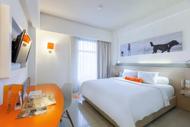 A hotel bedroom with a king bed, wall-mounted television, orange desk and chair, and a large beach photo of dogs above the headboard.