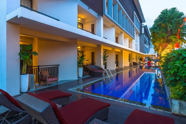 A long blue-tiled swimming pool runs beside a wooden deck with sun loungers and a multi-storey hotel building with balconies at dusk.